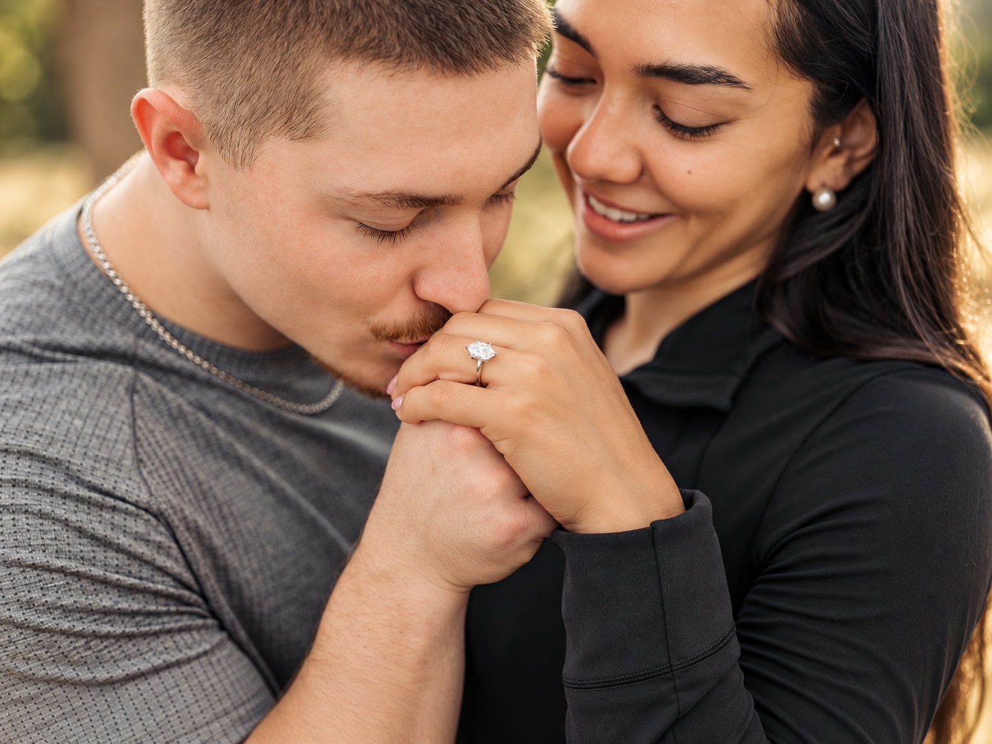 A couple sharing a hand-kiss moment outdoors, the woman's new engagement ring visible on her hand.
